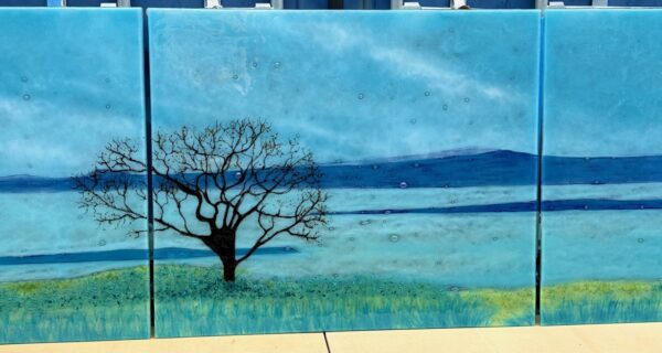 Three-panel fused glass artwork showing a lone tree beside blue estuary water and distant hills, measuring 200 x 91 cm.