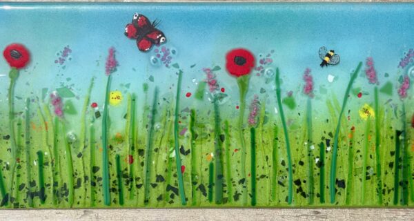 Fused glass wildflower meadow panel with red poppies, pink flowers, a butterfly and bee, measuring 52.6 x 20 cm.