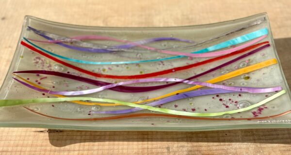 Handmade multicoloured fused glass bowl with flowing wave lines in red, turquoise, pink, purple, yellow and green, measuring 45 x 21 cm.